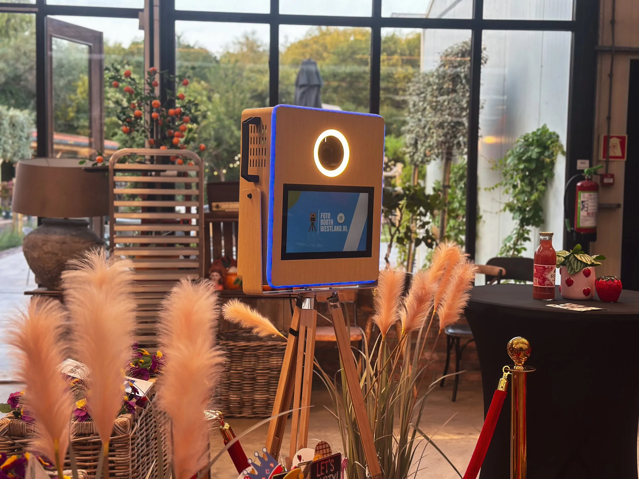 De photobooth op een houten statief in een lichte hal met grote ramen en pampasgras